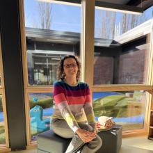 Image of woman with red glasses, brown hair, and striped sweater seated in a preschool classroom